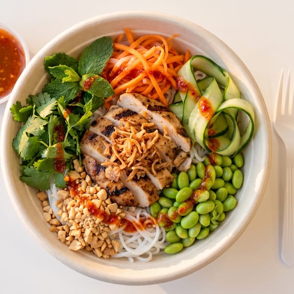 Grilled chicken, noodles, edamame, carrots, cucumber, and herbs in a bowl with sauce.